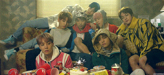 a gif from bts' mv spring day. it shows the 7 boys on a couch, watching a birthday cake with a lit sparkler-like candle.